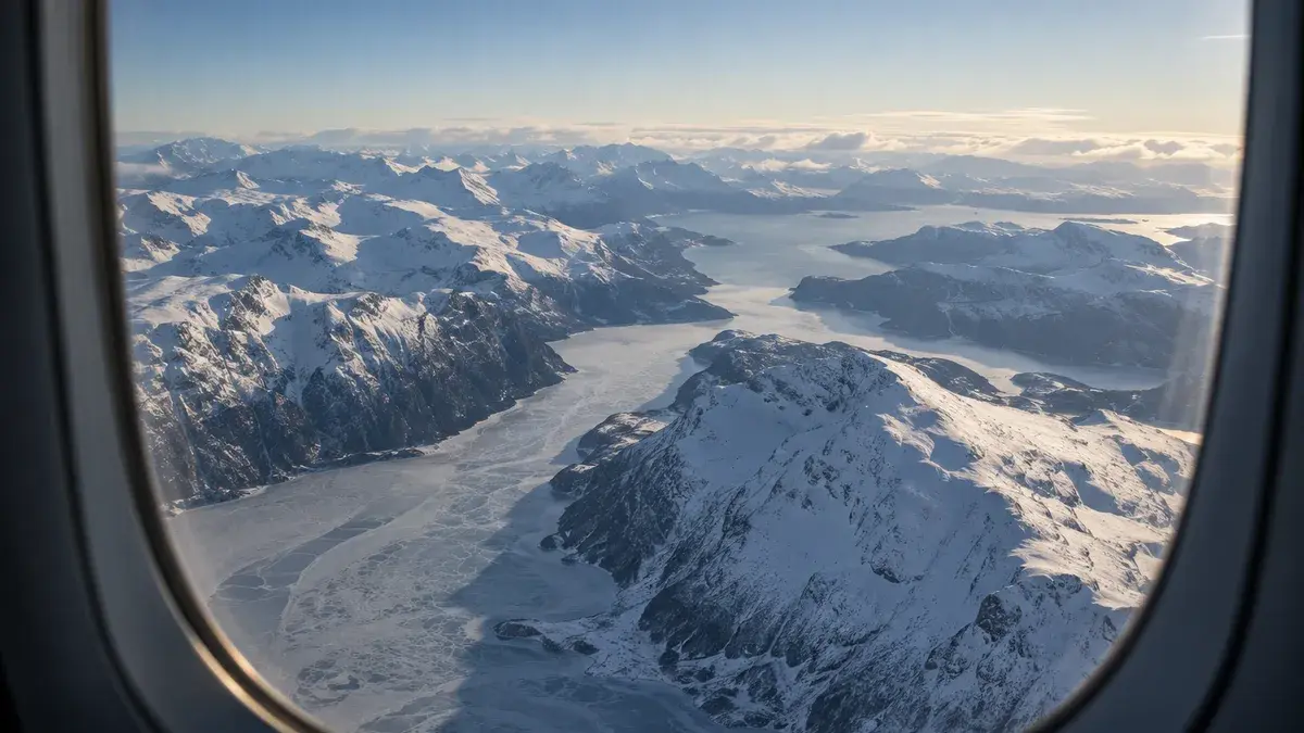 Utsikt gjennom et flyvindu over snødekte fjell og en islagt fjord på en klar vinterdag