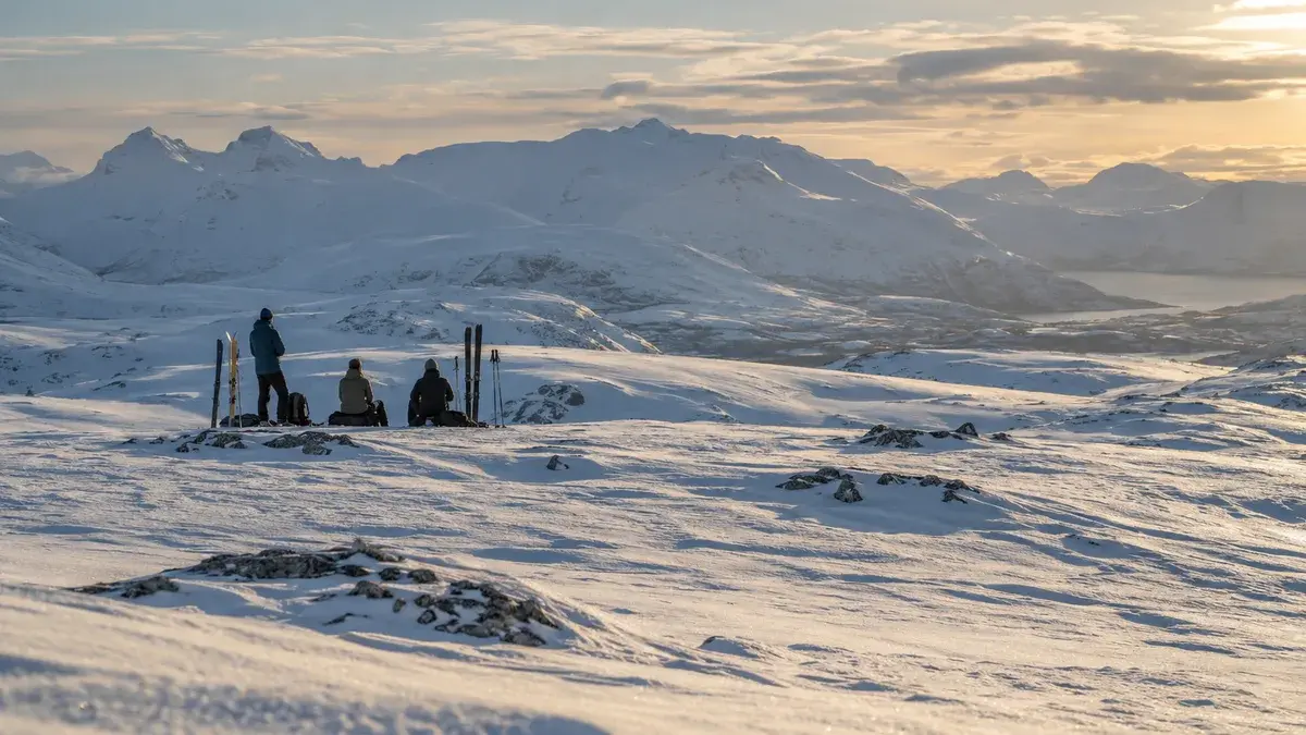 Tre skiløpere tar pause på et snødekt fjellplatå med ski stående i snøen og fjelltopper bak