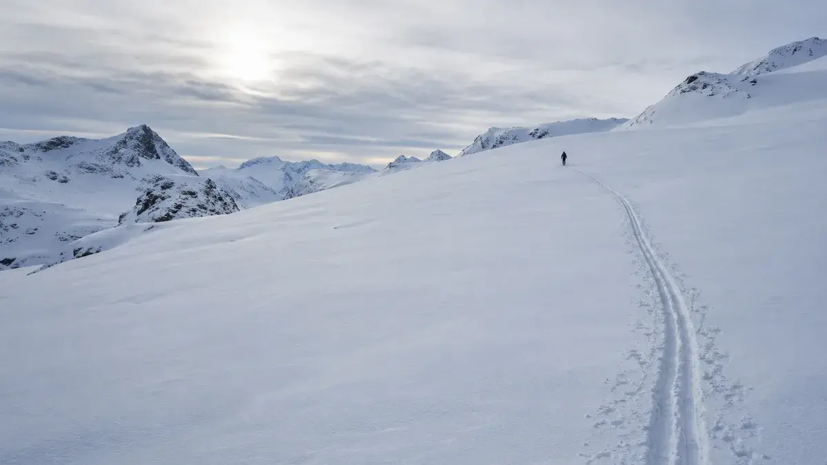 Et langt skispor svinger over en åpen snøbolle under tynt skydekke, med én liten skiløper langt oppe